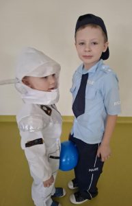 Dwóch chłopców stoi na przeciwko siebie w swoich przebraniach. Jeden z nich jest policjantem a drugi astronautą. Między ich nogami umieszczony jest niebieski balonik.