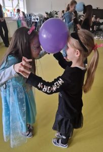 Dwie dziewczynki stoją na przeciwko siebie. Miedzy ich głowami umieszczony jest niebieski balon, który podtrzymują lekko pochylając głowy do przodu. Dziewczynki trzymają się za wyciągnięte w góre ręce.