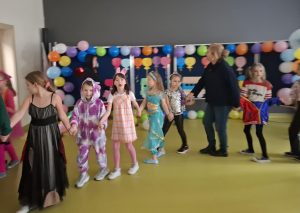 Uczniowie wraz z nauczycielką tańczą tworząc długiego węża, na sali gimnastycznej. Uczestnicy zabawy trzymają się za dłonie.