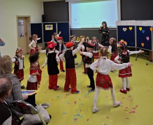 Uczniowie oddziału przedszkolnego tańczą tworząc swoimi ciałami dwa koła - w wewnętrznym kole stoją chłopcy z wycięgnietymi w górę prawymi rękami, a na zewnątrz dziewczynki odsuwając lewą rękę na zewnątrz koła. Na podłodze leży kolorowe konfetti a w tle widać stojącą wychowawczynię klasy.