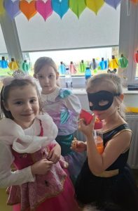 Trzy dziewczynki w swoich przebraniach na bal karnawałowy stoją obok siebie na sali teatralnej. w tle widać okno udekorowane kolorowymi serduszkami wyciętymi z papieru.