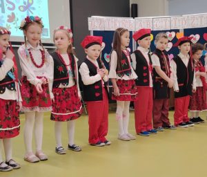Uczniowie oddziału przedszkolnego stoją w rzędzie w strojach krakowiaków. Dzieci patrzą przed siebie.