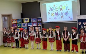 Uczniowie oddziału przedszkolnego w strojach krakowiaków stoją w jednym rzędzie. Za nimi na rzutniki wyświetla się napis: "KOCHAMY WAS".