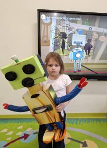 Uczennica oddziału przedszkolnego pozuje do zdjęcia trzymając w dłoniach przed sobą wykonaną na konkurs figurkę robota.Figurka prawie w całości zasłania dziewczynkę. W tle widoczny jest monitor, na którym wyświetlony jest obraz przedstawiający robota stojacego przed dwoma klatkami, w których umieszczone są czarne postaci przypominające ludzkie sylwetki.