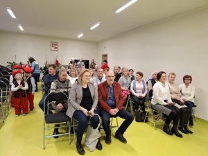 Dziadkowie i babcie uczniów oddziału przedszkolnego siedzą na krzesełkach ustawionych w rzędach na sali teatralnej. Obok drugiego rzędu stoją gęsiego dzieci w strojach krakowiaków.