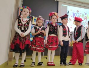 Trzy dziewczynki i dwóch chłopców stoi w rzędzie obok siebie. Dzieci ubrane są w stroje krakowiaków. Jeden z chłopców obraca w tył głowę.