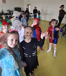 Uczniowie bawią się na sali teatralnej podczas balu karnawałowego. Dwie dziewczynki widoczne na pierwszym planie trzymają się za dłonie i machają w stronę obiektywu lekko uśmiechając się. Dzieci bawiące się w tle również machają dłońmi.