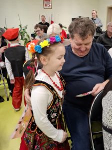 Uczennica oddziału przedszkolnego w stroju krakowianki stoi blisko obok swojej babci, która trzyma w dłoni otrzymaną laurkę. Obie patrzą w jej stronę. W tle widać innych uczniów i ich dziadków.