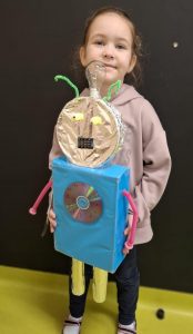 Uczennica oddziału przedszkolnego pozuje do zdjęcia trzymając przed sobą wykonanego na konkurs robota. dziewczynka uśmiecha się a robot zasłania połowę jej działa. Za uczennicą widoczna jest czarna ściana.