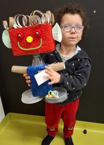 Uczennica oddziału przedszkolnego pozuje do zdjęcia trzymając przed sobą wykonanego na konkurs robota. Głowa robota znajduje się na wysokości twarzy dziewczynki. Zdjęcie wykonane na tle czarnej ściany.