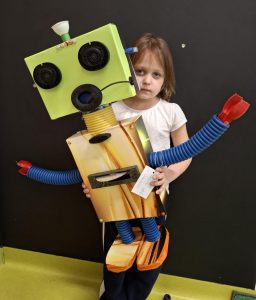 Uczennica oddziału przedszkolnego pozuje do zdjęcia trzymając przed sobą wykonanego na konkurs robota. Figurka prawie w całości przesłania postać dziewczynki. Zdjęcie wykonane na tle czarnej ściany.