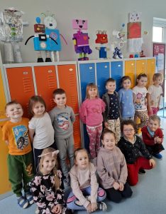 Uczniowie oddziału przedszkolnego pozują do wspólnego zdjęcia na szkolny korytarzu. Za dziećmi widoczne są kolorowe szafki, na których umieszczone zostały figurki robotów wykonane przez dzieci. Część z uczniów stoi opartych plecami o szafki, a przed nimi na podłodze siedzą ich koledzy.