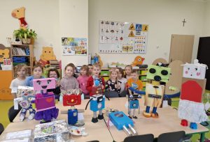 Uczniowie oddziału przedszkolnego stoją w grupie za dużym stołem. Na stole ustawione są wykonane przez uczniów figurki robotów.