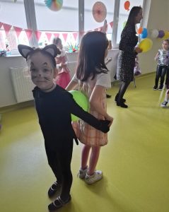 Dwie uczennice w swoich przebraniach karnawałowych stoi do siebie plecami trzymając się za dłonie. Pomiędzy dziewczynkami umieszczony jest jasnozielony balon. Dziewczynka w stroju kota zwrócona jest przodem w stronę obiektywu.