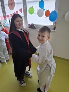 Dwoje uczniów tańczy podczas balu karnawałowego trzymając się za ręce. Dziewczynka ubrana jest w czarną szatę ozdobioną czerwonym krawatem, a chłopiec jest ubrany na biało w strój dżudoki.