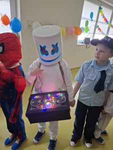 W centralnej części zdjęcia widoczny jest chłopiec ubrany na biało, którego głowa zasłonięta jest zakryciem w kształcie walca z wyciętymi otworami na oczy i namalowanym czarnym szerokim uśmiechem. Zarówno brzegi wycięć na oczy jak i linia uśmiechu podświetlona jest na niebiesko. Uczeń ma zawieszone na szyi czarne pudełko z podświetlonym kolorowymi lampkami zdjęciem konsoli djeskiej.