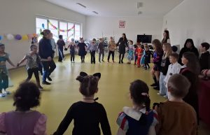 Uczniowie wraz ze swoimi wychowawczyniami tańczą na balu. Wszyscy tworzą duże koło trzymając za ręce swoich sąsiadów.