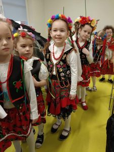 Uczennice w strojach krakowianek z kolorowymi wiankami na głowach stoją w rzędzie jedna za drugą. Część dziewczynek się wychyla i uśmiecha.