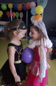 Dwie dziewczynki stroją blisko obok siebie zwrócone twarzami w swoim kierunku. Na wysokości brzucha umieszczony mają balon, który wspólnie podtrzymują swoimi ciałami.