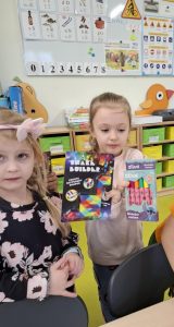 Uczennica oddziału przedszkolnego pozuje do zdjęcia trzymając przed sobą w dłoniach dwie nagrody - pudełko z pisakami w prawej dłoni i drugie z łamigłówką matematyczna w lewej. Obok stoi jej koleżanka.