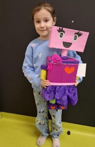 Uczennica oddziału przedszkolnego pozuje do zdjęcia trzymajac w dłoniach przed sobą wykonanego na konkurs robota. Robot zasłania część sylwetki dziewczynki, a jego głowa kończy się na wysokości jej ust. Zdjęcie wykonane na tle czarnej ściany.