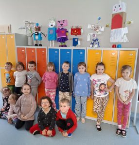 Uczniowie oddziału przedszkolnego pozują do wspólnego zdjęcia przy kolorowych szkolnych szafkach, na których ustawione są przygotowane przez uczniów figurki robotów. Część uczniów stoi przy szafkach oparte o nie plecami. Przed nimi na podłodze siedzą ich koledzy.