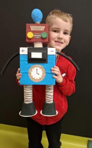 Uczeń oddziału przedszkolnego pozuje do zdjęcia trzymając w dłoniach przed sobą wykonanego na konkurs robota. Figurka zasłania tułów chłopca, który swoja głowę przyciska do głowy robota. Chłopiec uśmiecha się pokazując zęby. Zdjęcie wykonane na tle czarnej ściany.