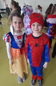 Dwoje uczniów pozuje do wspólnego zdjęcia podczas balu karnawałowego. Dziewczynka ubrana jest w strój księżniczki a chłopiec przebrany jest za spider-mena. Dzieci uśmiechają się. W tle widać inne dzieci, które tańczą.