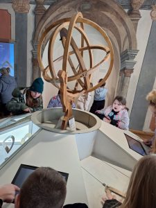 W centralnym miejscu zdjęcia widoczna jest konsola z monitorami umieszczonymi na platformie pośrodku, której widoczna jest drewniana rzeźba przedstawiająca postać trzymającą w dłoniach krzyżujące się okręgi umieszczone wewnątrz siebie. Wokół konsoli stoją uczniowie wpatrzeni w monitor. Jedna z dziewczynek patrzy z otwartą buzią w kierunku rzeźby.