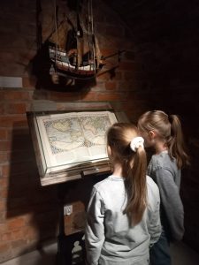 Dwie uczennice stoją przed wielką otwartą księgą, na której stronach widać mapę kartograficzną. Nad książką przy ścianie z cegieł wisi model statku. Dziewczynki odwrócone są plecami do obiektywu.