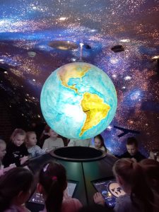 Uczniowie biorący udział w wyjeździe stoją wokół czarnej konsoli z umieszczonymi w niej ekranami. Na środku nad konsolą unosi się podświetlony model kuli ziemskiej. Dzieci patrzą na ekrany konsoli. W tle widoczna jest ściana z podświetlonymi punktami świetlnymi, której malowanie replikuje nocne niebo z widocznymi pasmami gwiazd.