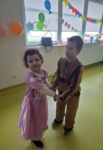 Dwoje uczniów oddziału przedszkolnego tańczy wspólnie trzymając się za dłonie. Dziewczynka ubrana jest w strój księżniczki a chłopiec indianina. Dziewczynka lekko odchyla głowę w stronę obiektywu uśmiechając się.