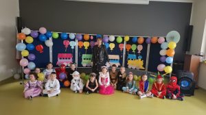 Uczniowie oddziału przedszkolnego siedzą obok siebie, w swoich kostiumach na podłodze sali teatralnej. za nimi stoi ich wychowawczyni. Za nią widoczna jest tablica ozdobiona kolorowymi, nadmuchanymi balonami oraz wyciętym z kolorowego papieru pociągiem.