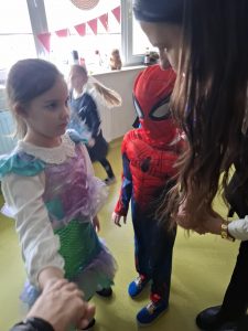 Zdjęcie przedstawia panią nauczycielkę pochylającą się nisko do stojącej na przeciwko dziewczynki ubranej w strój syrenki. Obok stoi chłopiec przebrany za Spider-Mena.