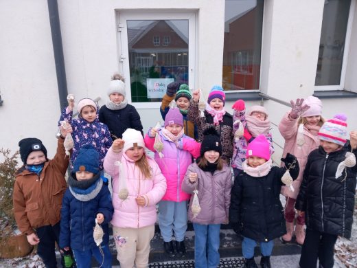Uczniowie klasy pierwszej stoją ubrani w kurki i czapki przed bocznym wejściem do szkoły. Dzieci trzymają w wyciągniętych przed soba dłoniach woreczki z pokarmem dla ptaków.