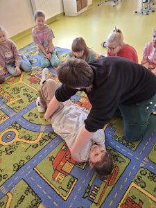 Na dywanie leży uczennica ze splecionymi wokół ciała swoimi rękoma. Nad nią pochyla się Pani prowadząca szkolenia trzymająca dłonie tak by przewrócić jej ciało do pozycji ustalonej bocznej. Całą scenę obserwują inni uczniowie siedzący w bliskiej odległości na dywanie.