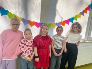 Pięć uczennic stoi w rzędzie przy oknie na którym została zawieszona girlanda z kolorowych serduszek. Dwie z dziewczynek mają założone na twarzy różowe okulary w kształcie serc.