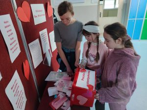 Trzy uczennice stoją przy stoliku, na który wysypują karteczki z czerwonego pudełka. Pudełko ozdobione zostało wyciętymi z papieru serduszkami oraz kartką z napisem "Poczta walentynkowa".