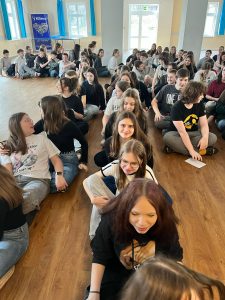 Uczniowie biorący udział w warsztatach siedzą na podłodze w rzędach , które pod koniec zawracają. Na pierwszym planie widoczni są uczniowie naszej szkoły siedzący w prostym rzędzie jeden za drugim.