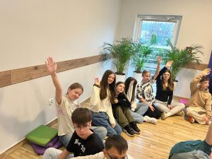 Uczniowie naszej szkoły biorący udział w wyjeździe siedzą na podłodze. Część z nich wysoko podnosi w górę jedną ze swoich rąk.