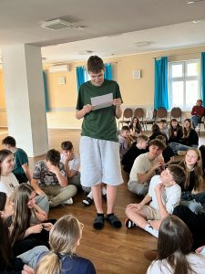 Uczeń klasy ósmej stoi na środku sali patrząc na kartkę papieru trzymającą w dłoniach przed sobą. Wokół niego na podłodze siedzą inni uczestnicy warsztatów.