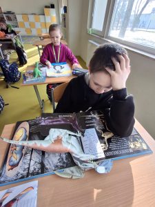 Uczeń klasy drugiej siedzi przy stole. Przed nim leży otwarta książka z informacjami o dinozaurach. Chłopiec jedną z rąk opiera o książkę a jego dłoń podpiera pochyloną w jej stronę głowę. W tle widać siedzącą przy kolejnym stoliku uczennicę.