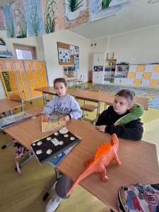 Dwie uczennice siedzą przy stoliku,, na którym leżą książki oraz duża figurka dinozaura. Jedna z dziewczynek przytula dużą zieloną maskotkę.