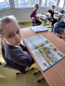 Uczennica klasy drugiej siedzi przy stoliku. Dziewczynka patrzy w stronę obiektywu, a przed nią leży otwarta książka z dinozaurami. W tle widać siedzących przy swoich stolikach innych uczniów.