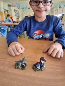 Uczeń klasy drugiej siedzi przy stoliku na którym opiera swoje dłonie. Przed chłopcem stoją dwie małe figurki przedstawiające dinozaury.
