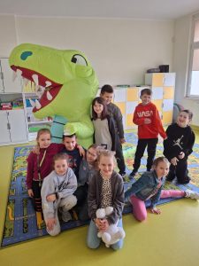 Uczniowie klasy drugiej pozują do wspólnego zdjęcia z osoba przebraną za dinozaura. Część dzieci kuca, a inne stoją i przytulają się do maskotki. Uczniowie są uśmiechnięci. Dinozaur stoi bokiem.
