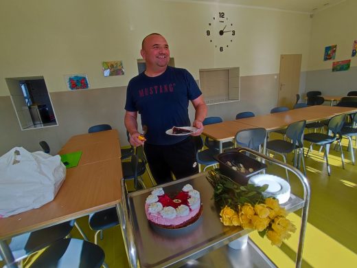 Nauczyciel szeroko uśmiecha się stojąc w szkolnej stołówce. Mężczyzna w jednej dłoni trzyma nóż, a w drugiej, talerz z kawałkiem tortu. Przed nim na srebrnej tacy ułożony jest tort oraz pęk żółtych róż.