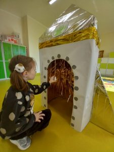 Uczennica klasy pierwszej kuca przed wykonaną z papieru i ozdobioną folia aluminiową repliką rakiety kosmicznej. Dziewczynka dotyka dłonią przedmiotu zawieszonego na sznurku przy otworze pozwalającym wejść do rakiety.