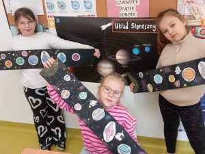 Trzy uczennice pozują do wspólnego zdjęcia trzymając długie paski czarnego papieru ozdobione rysunkami i naklejkami planet i obiektów związanych z przestrzenią kosmiczną. Dziewczynki stoją na tle ściany, na której zawieszony jest plakat zatytułowany - "Układ Słoneczny".