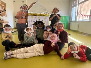 Uczniowie klasy pierwszej pozują do wspólnego zdjęcia. Dzieci na wysokości twarzy trzymają ramki wycięte z papieru w kształcie hełmów astronautów. Za nimi stoi makieta rakiety kosmicznej ozdobiona folią aluminiową.